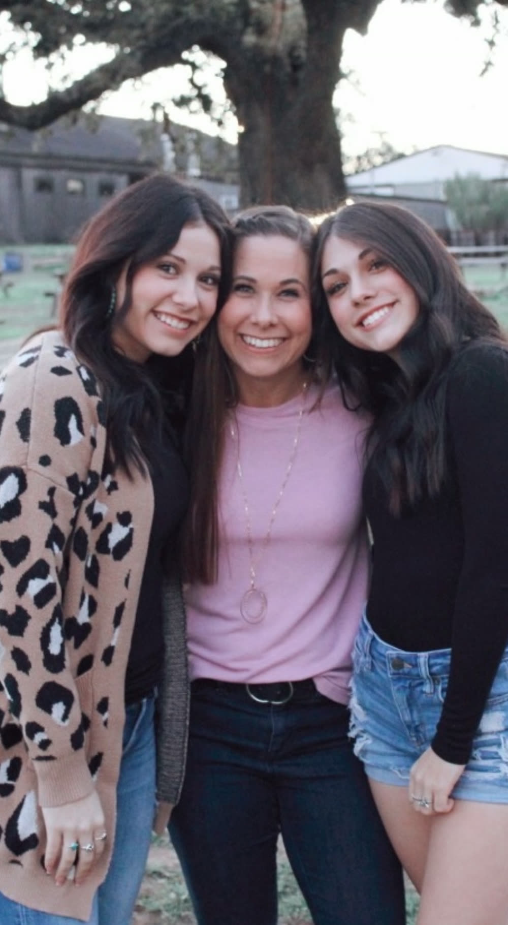 Carly standing outdoors under an oak tree between her two daughters, all three smiling.